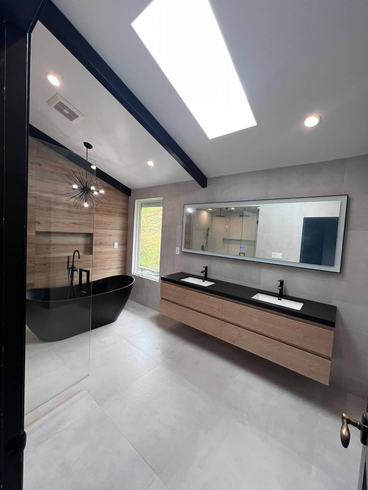 Bathroom remodel with black freestanding tub, floating wood vanity, skylight, and wide mirror