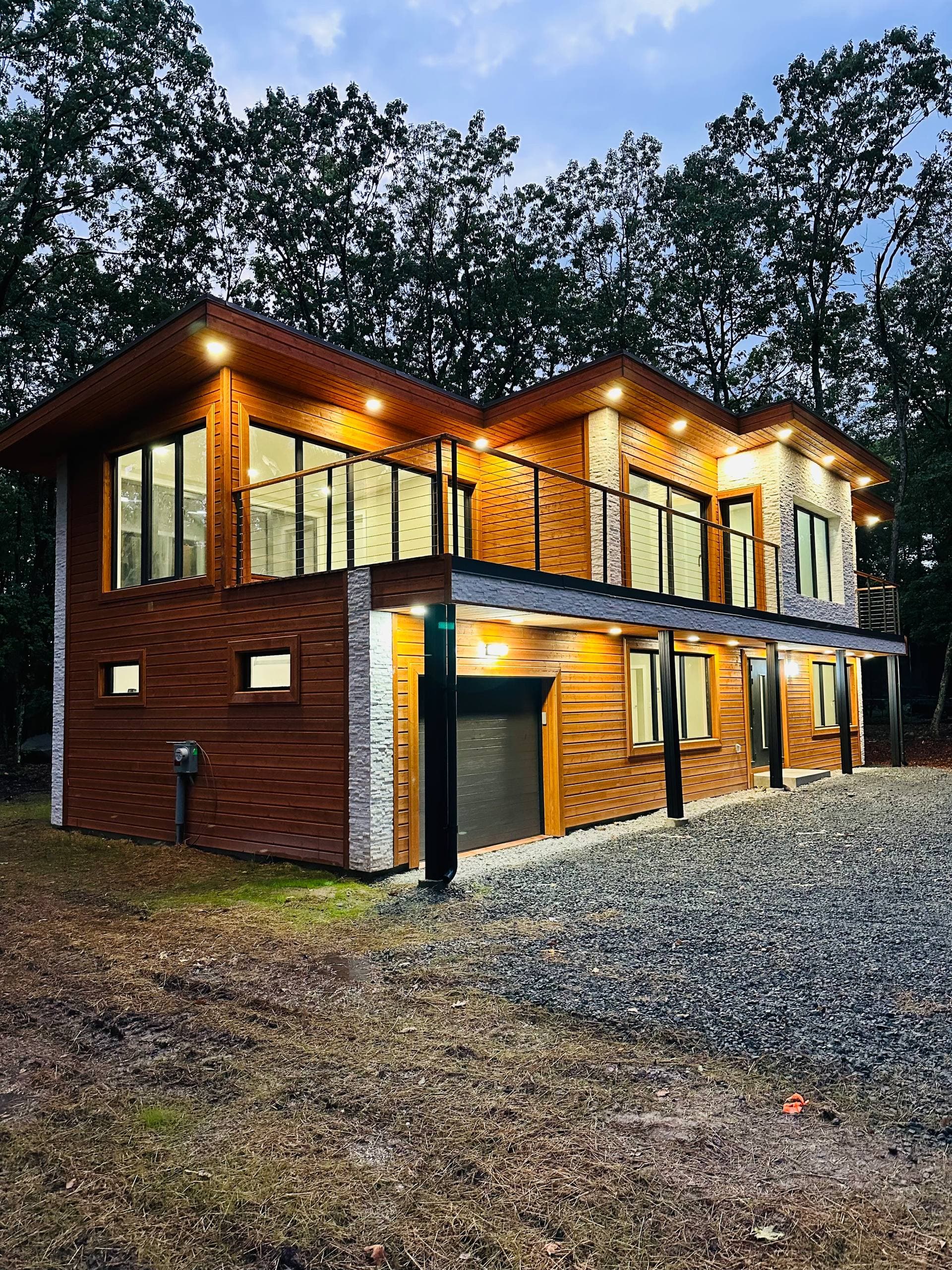 Modern wood exterior home with balcony, black railings, stone accents, and evening lighting